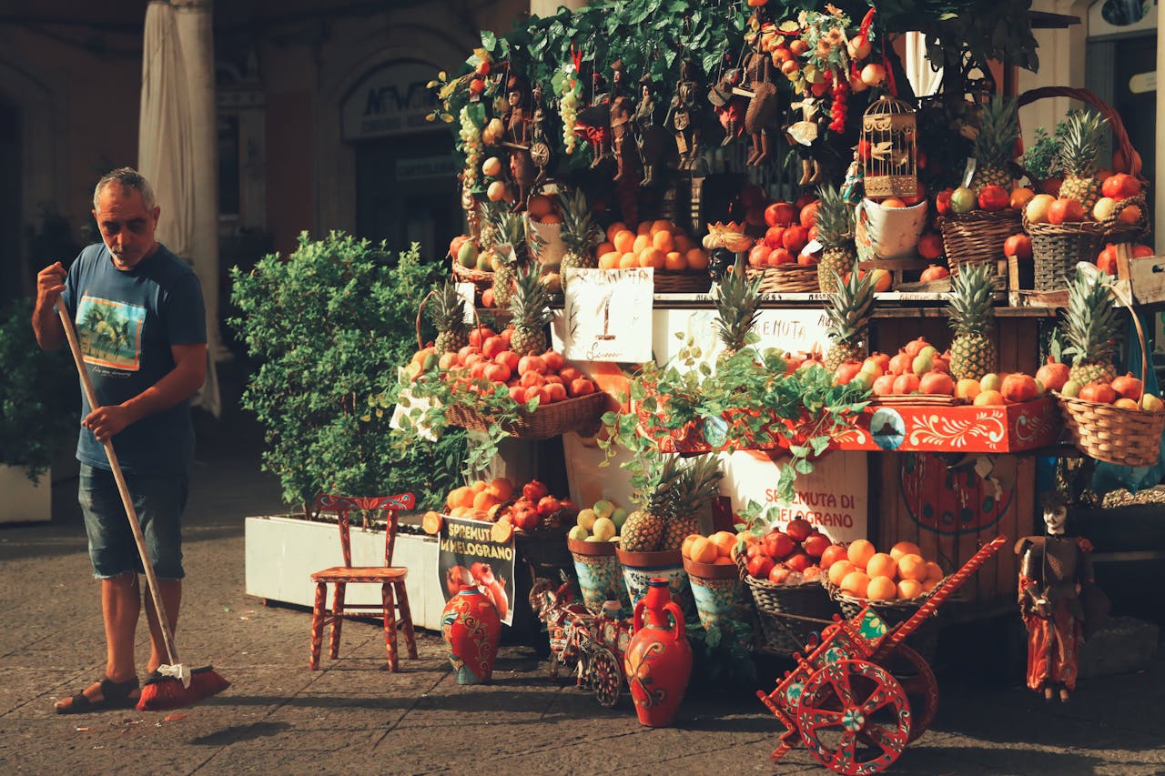 A man sweeps next to a colorful fruit stand in Catania's bustling market, Sicily, Italy.